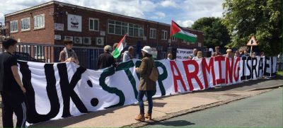 Protesters in front of a factory hold up a banner saying "UK: Stop Arming Israel"